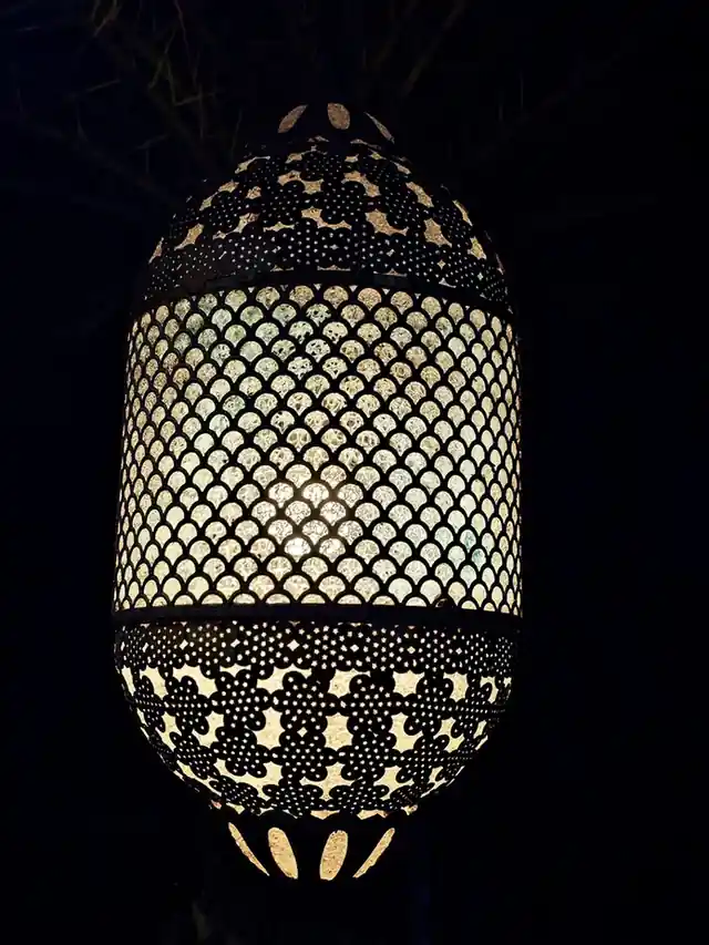A tall pierced metal lantern glowing against a dark room, its scale-pattern holes and cut-out band throwing pinpoints of light